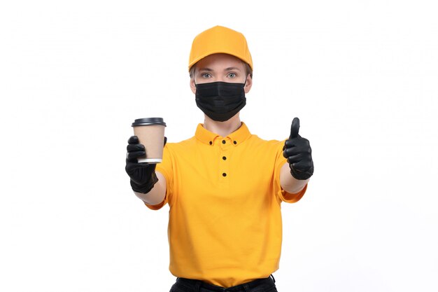 A front view young female courier in yellow uniform black gloves and black mask holding coffee cup