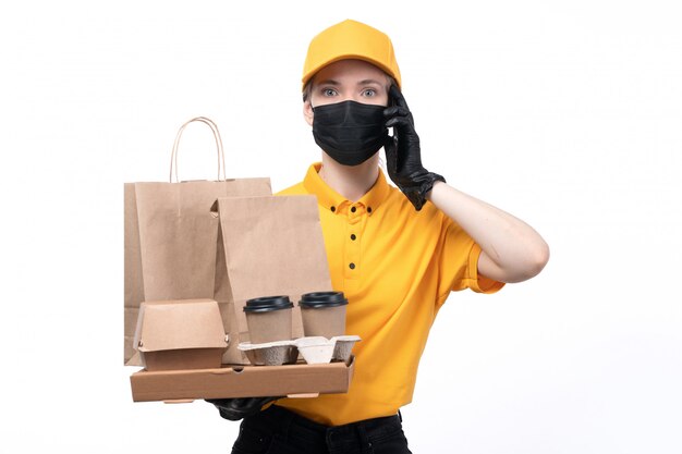 Молодая женщина-курьер в желтой униформе, черные перчатки и черная маска, держащая кофейные чашки, разговаривает по телефону