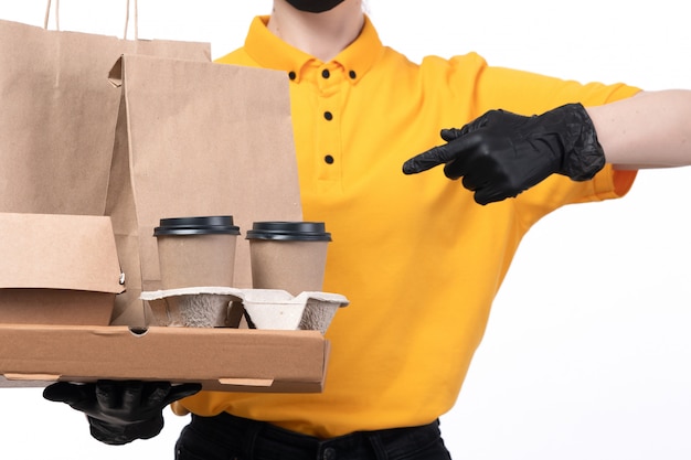 Вид спереди молодая женщина-курьер в желтой униформе, черные перчатки и черная маска, держащая пакеты для доставки еды из кофейных чашек