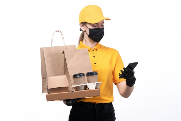 A front view young female courier in yellow uniform black gloves and black mask holding coffee cups food delivery packages