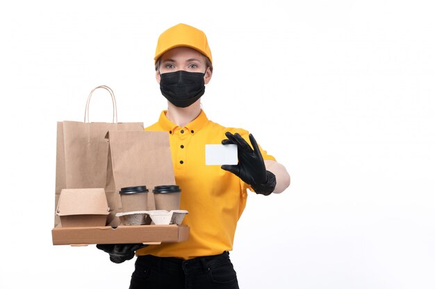 A front view young female courier in yellow uniform black gloves and black mask holding coffee cups and food packages