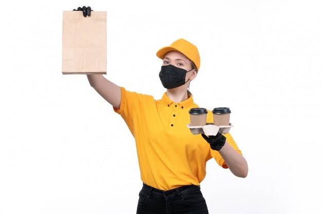 A front view young female courier in yellow uniform black gloves and black mask holding coffee cups and package