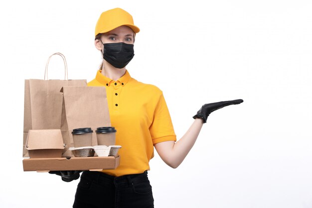 A front view young female courier in yellow uniform black gloves and black mask holding coffee cups and packages