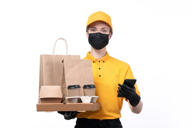 黄色の制服の黒い手袋とコーヒーカップの電話とパッケージを保持している黒いマスクの正面の若い女性の宅配便