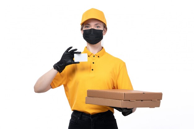 Молодая женщина-курьер в желтой униформе, черные перчатки и черная маска, держащая пакеты для доставки еды и белую карточку, вид спереди