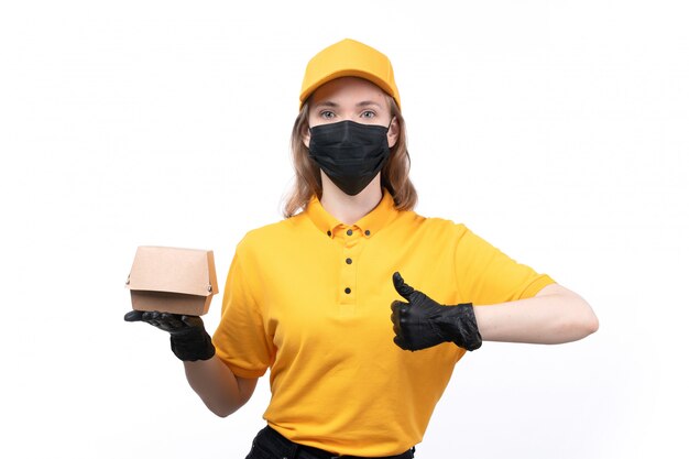 A front view young female courier in yellow uniform black gloves and black mask holding food package showing like sign