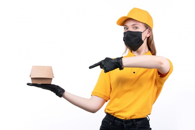 A front view young female courier in yellow uniform black gloves and black mask holding food package