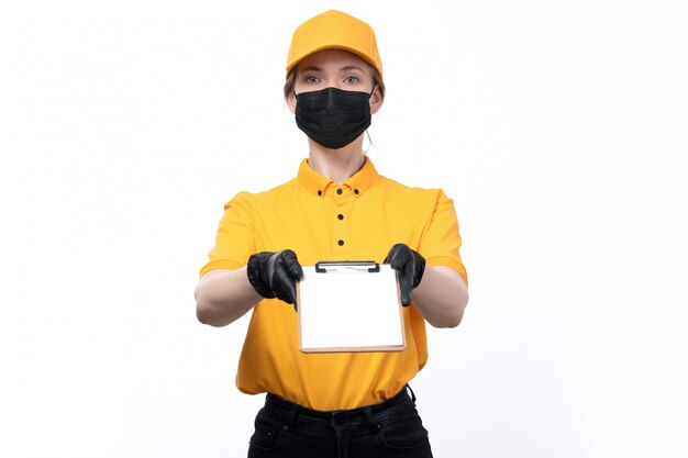 A front view young female courier in yellow uniform black gloves and black mask holding notepad for signature