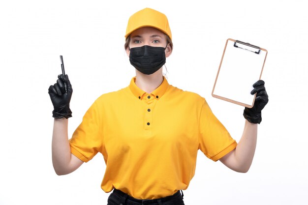 A front view young female courier in yellow uniform black gloves and black mask holding notepad