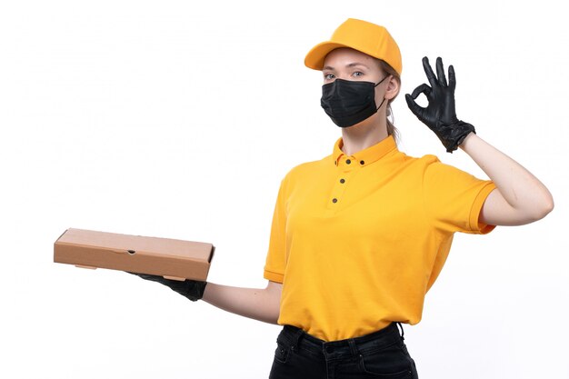 A front view young female courier in yellow uniform black gloves and black mask holding pizza box showing alright sign