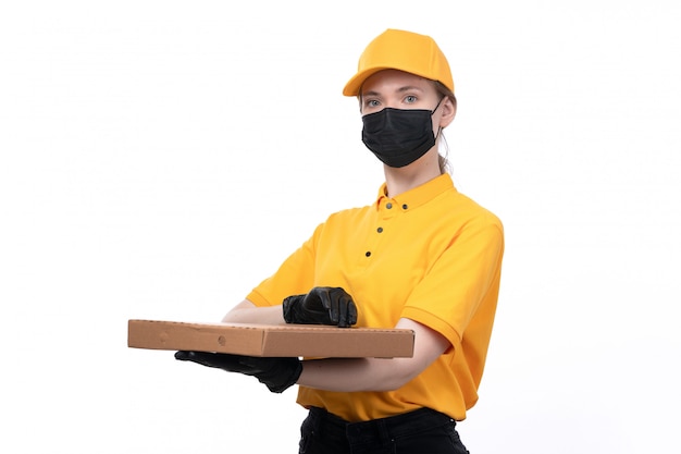 A front view young female courier in yellow uniform black gloves and black mask holding pizza box