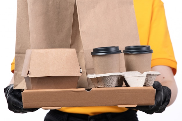 A front view young female courier in yellow uniform black gloves and black mask holding pizza boxes and coffee cups