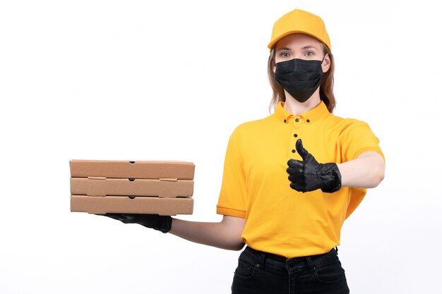 A front view young female courier in yellow uniform black gloves and black mask holding pizza boxes showing like sign