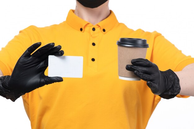 A front view young female courier in yellow uniform black gloves and black mask holding white card and coffee cup