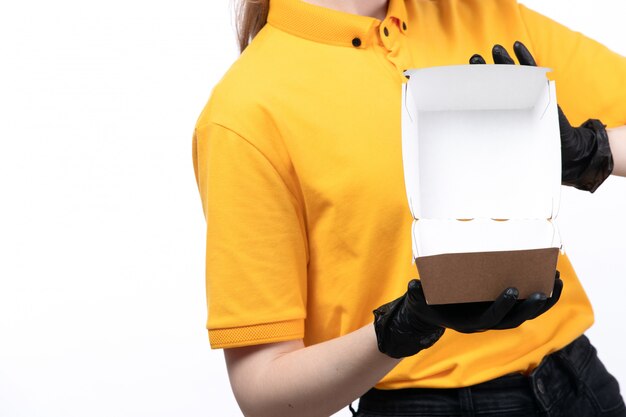 A front view young female courier in yellow uniform black glvoes holding an empty food package