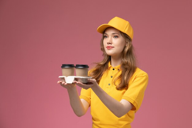 Вид спереди молодая женщина-курьер в желтой форме, доставляющая кофе на розовом столе, работник службы доставки униформы