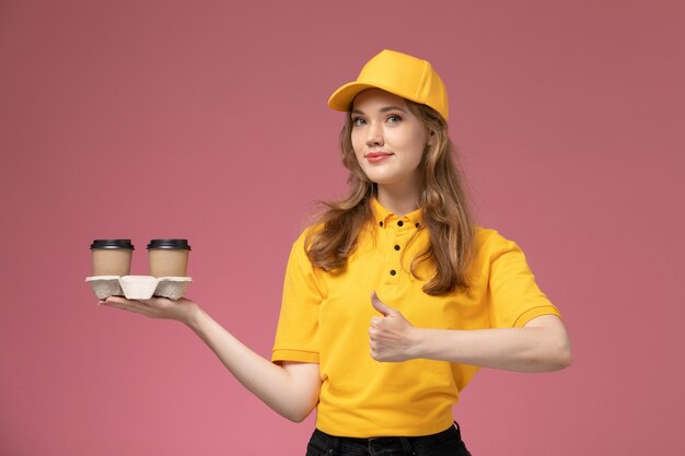 Front view young female courier in yellow uniform holding brown coffee cups on the pink desk job uniform delivery service worker