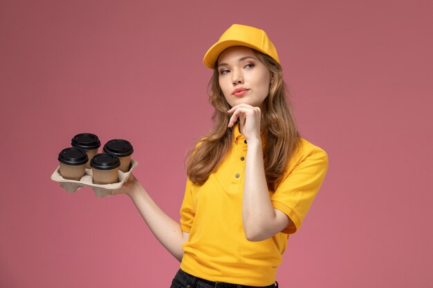 濃いピンクのデスクの制服配達サービスの女性労働者を考えて茶色のコーヒープラスチックカップを保持している黄色の制服を着た若い女性の宅配便の正面図