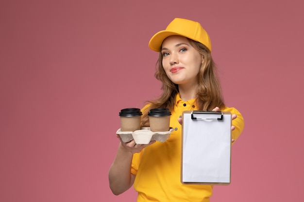 Front view young female courier in yellow uniform holding brown coffee plastic cups with notepad on dark-pink desk uniform delivery service female worker