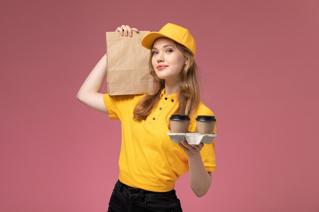 Вид спереди молодая женщина-курьер в желтой форме, держащая кофейные чашки и пакет с едой на розовом фоне, рабочий стол службы доставки униформы