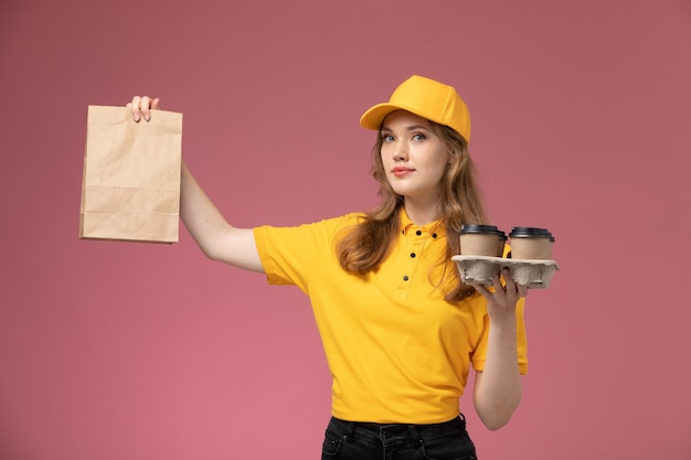 분홍색 책상 책상 작업 유니폼 배달 서비스 노동자에 음식과 커피 컵과 패키지를 들고 노란색 제복을 입은 전면보기 젊은 여성 택배