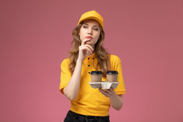 Front view young female courier in yellow uniform holding coffee cups posing with thinking expression on dark pink desk uniform delivery job service worker
