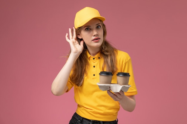 Free photo front view young female courier in yellow uniform holding coffee cups trying to hear out on dark pink desk uniform delivery job service worker