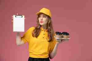 Free photo front view young female courier in yellow uniform holding delivery coffee and notepad posing on the pink desk job uniform delivery service worker