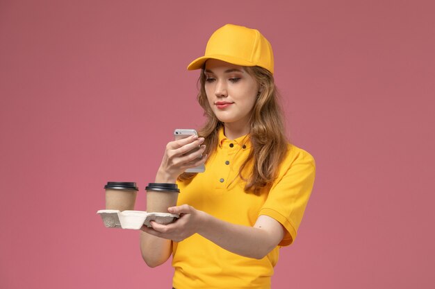 Front view young female courier in yellow uniform holding delivery coffee and using her phone on the pink desk job uniform delivery service worker