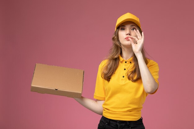 Front view young female courier in yellow uniform holding delivery food box and posing on pink desk job uniform delivery service worker