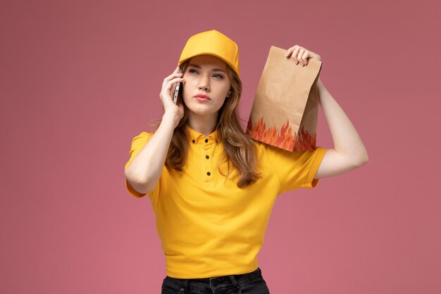 Front view young female courier in yellow uniform holding delivery food package and talking on the phone on pink desk job uniform delivery service worker