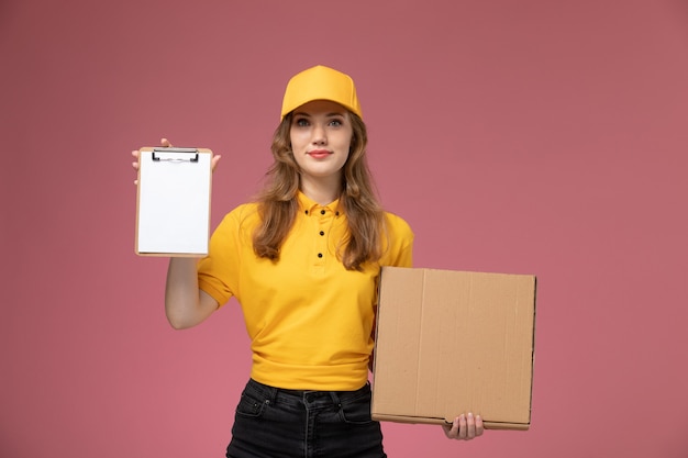 Вид спереди молодая женщина-курьер в желтой форме, держащая коробку для еды с блокнотом на темно-розовом фоне, работник службы доставки униформы