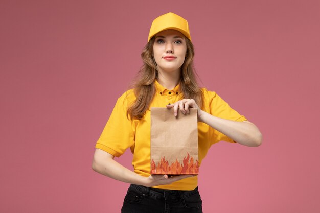 ピンクの背景の仕事の制服の配達サービスでそれを配達する食品パッケージを保持している黄色の制服を着た若い女性の宅配便の正面図