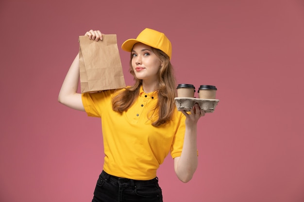 Вид спереди молодая женщина-курьер в желтой форме, держащая пакет с едой и кофейными чашками на розовом фоне, рабочий стол службы доставки униформы