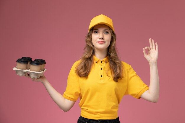 Вид спереди молодая женщина-курьер в желтой униформе держит пластиковые коричневые кофейные чашки с милым выражением лица на розовом фоне.