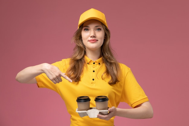분홍색 배경 작업 유니폼 배달 색상 서비스 노동자에 그녀의 얼굴에 미소로 플라스틱 갈색 커피 컵을 들고 노란색 제복을 입은 전면보기 젊은 여성 택배
