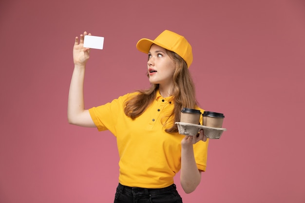 Вид спереди молодая женщина-курьер в желтой форме, держащая пластиковую кофейную чашку с карточкой на темно-розовом столе, работник службы доставки униформы