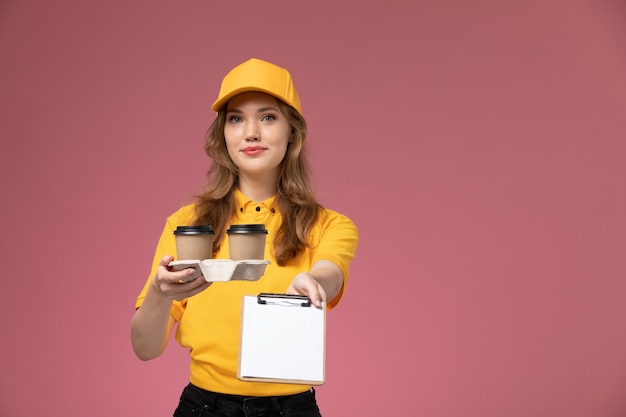 Giovane corriere femminile di vista frontale in uniforme gialla che tiene tazze di caffè di plastica e blocco note sul lavoratore di servizio di consegna uniforme scrivania rosa scuro