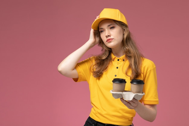 プラスチック製のコーヒーカップを保持し、濃いピンクの背景の制服配達ジョブサービスワーカーについて考える黄色の制服を着た若い女性の宅配便の正面図