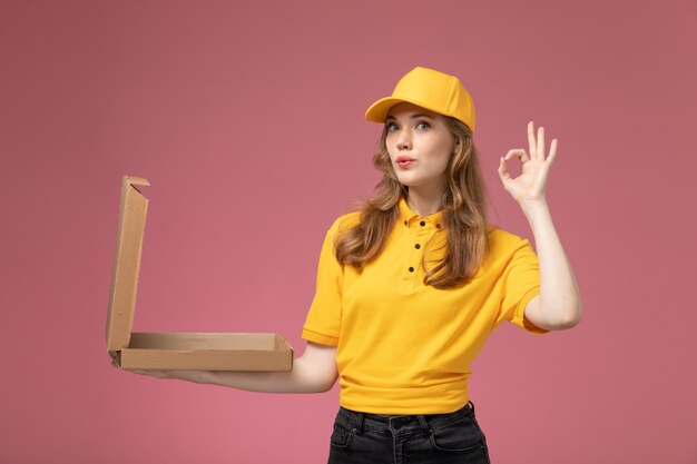 ピンクの机の上の開いたフードボックスでポーズをとって黄色の制服を着た若い女性の宅配便の正面図仕事制服配達サービスワーカー