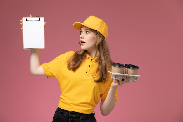 Front view young female courier in yellow uniform yellow cape holding coffee cups and notepad on the dark-pink background uniform delivery job service color