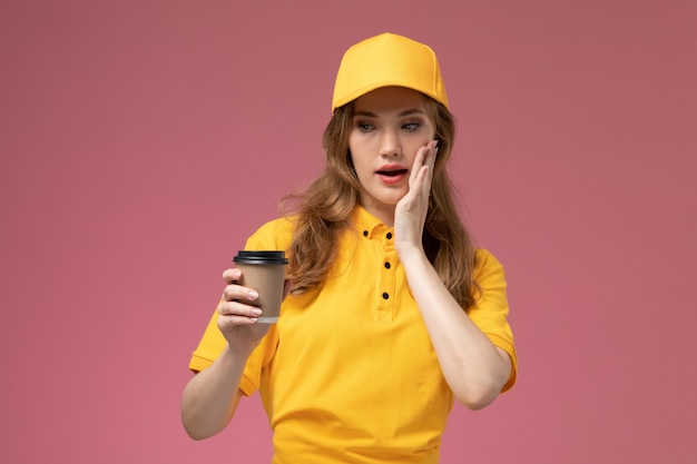 Front view young female courier in yellow uniform yellow cape holding coffee on dark-pink background uniform delivery service color