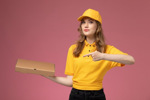 Front view young female courier in yellow uniform yellow cape holding food delivery box on the dark-pink background uniform delivery job service color