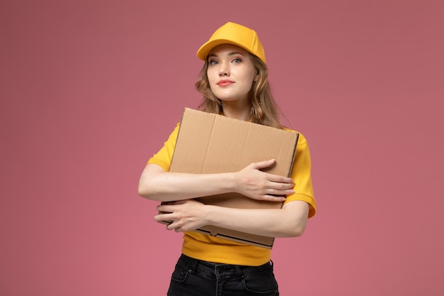 Corriere femminile giovane vista frontale in mantello giallo uniforme giallo che tiene la scatola di consegna del cibo sul colore di servizio di lavoro di consegna uniforme sfondo rosa scuro