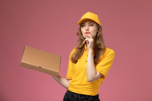 Front view young female courier in yellow uniform yellow cape holding food delivery box on the dark pink background uniform delivery job service color