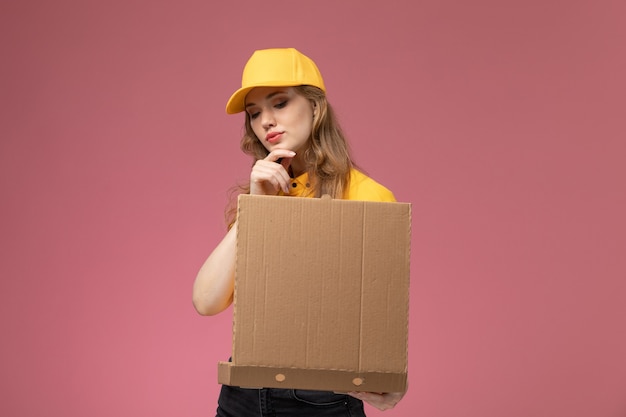 Front view young female courier in yellow uniform yellow cape holding food delivery box on the dark-pink desk uniform delivery job service color