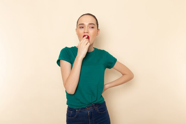 Front view young female in dark green shirt and blue jeans biting fruit on beige