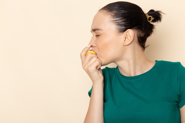 Front view young female in dark green shirt and blue jeans biting sour lemon on beige