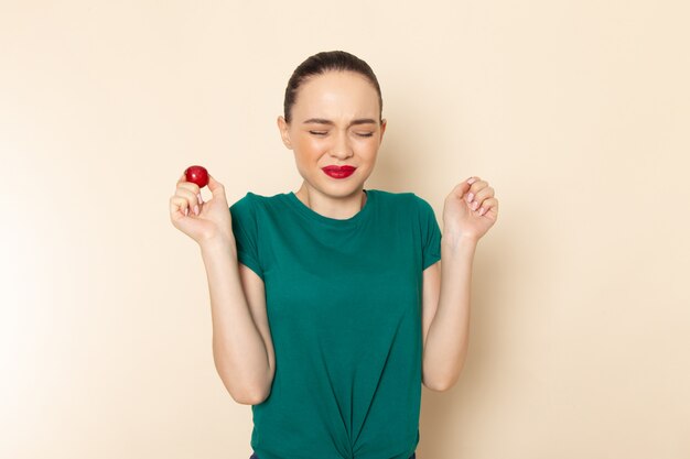 Front view young female in dark green shirt and blue jeans biting sour plum on beige