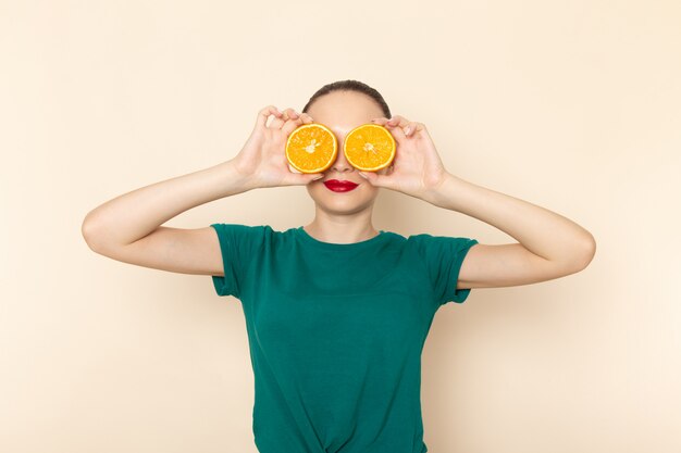 Front view young female in dark green shirt and blue jeans covering her eyes with oranges on grey